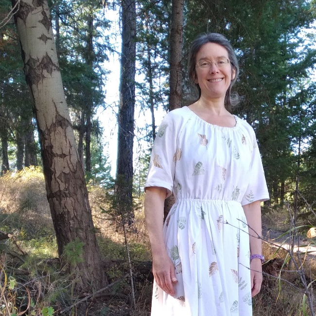 Middle aged white female with glasses and a white dress, standing in the woods with sunshine behind. 